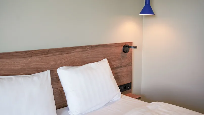 A neatly made bed with white pillows rests against a wooden headboard. A blue wall lamp is mounted above, softly illuminating the minimalist bedroom.