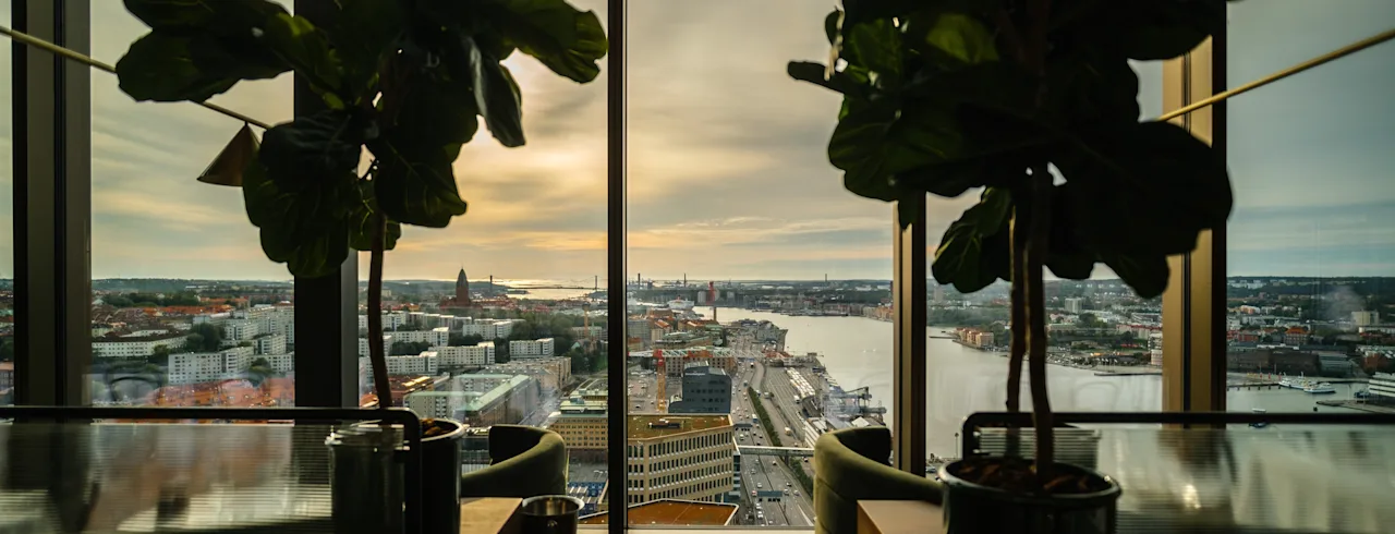 A high-rise view of a city and a river at sunrise or sunset, framed by large potted plants in a modern lounge area.