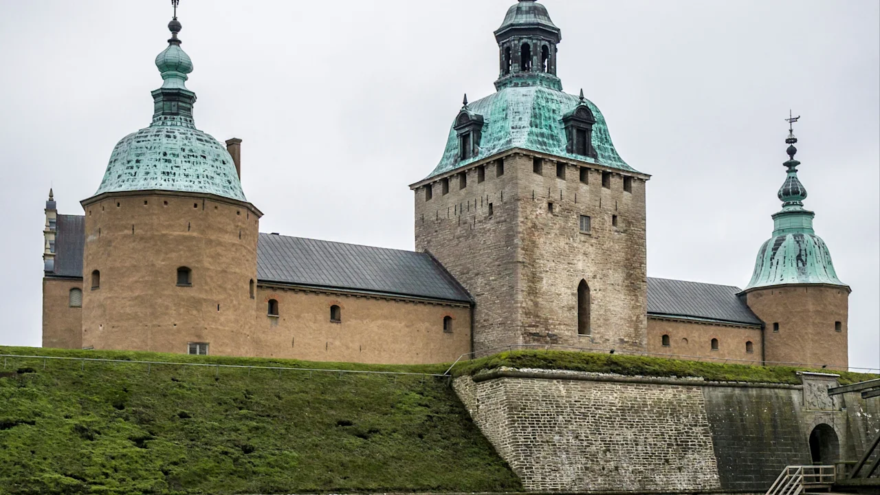 Et historisk stenborg med tre grønne kobbertårne står på græsklædte bredder, omgivet af en voldgrav under en skyet himmel.