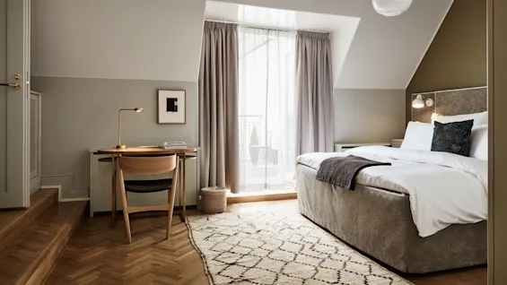 Et koselig soverom har en pent oppredd dobbeltseng med hvitt sengetøy, et skrivebord med lampe og stol i nærheten, og mykt naturlig lys som filtreres gjennom gardiner.