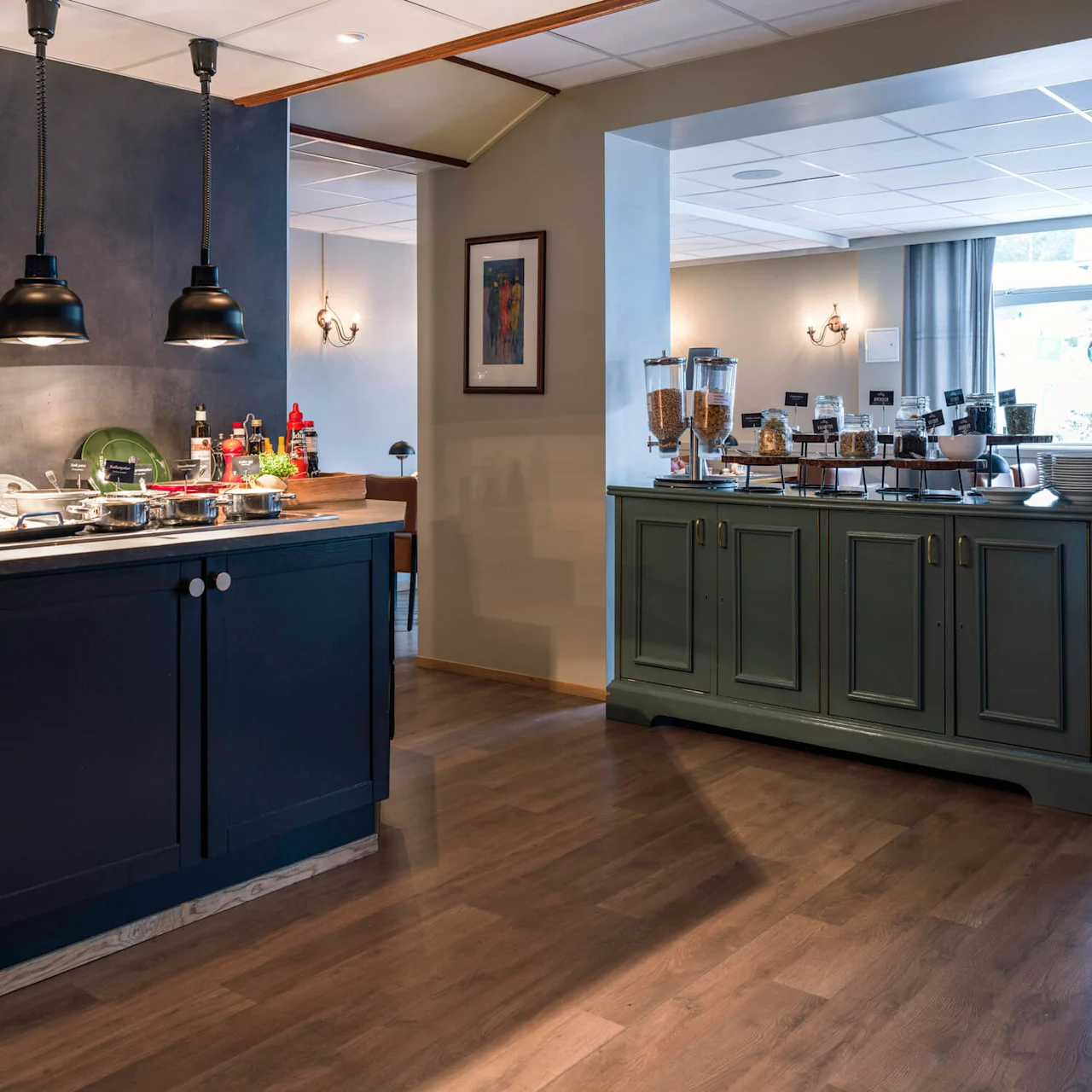 Kitchen counter displays cooking equipment and colorful dishes under pendant lights; adjacent buffet counter holds cereal dispensers and bowls, situated in a modern dining space with wooden flooring.