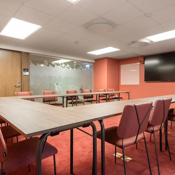 A large U-shaped conference table is surrounded by red chairs in a well-lit meeting room with red carpet, beige walls, a wooden door, and a mounted flat-screen TV.
