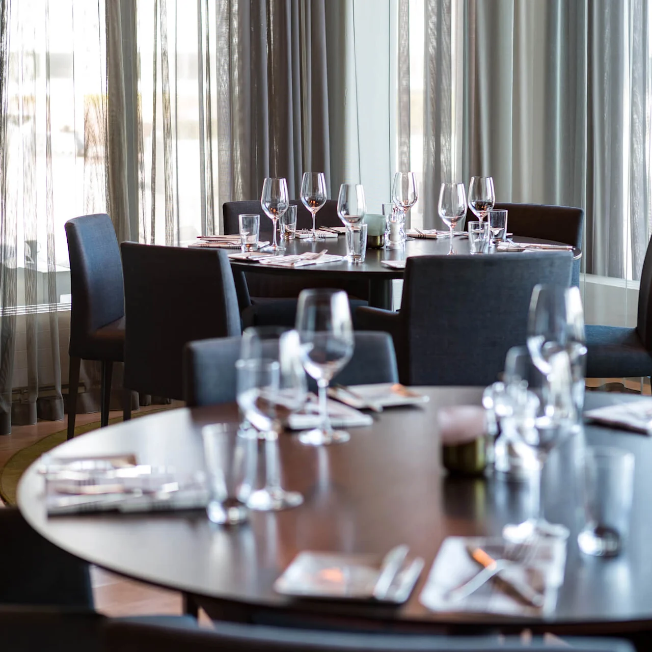 Tables set with glassware and cutlery, surrounded by chairs. Hanging lights illuminate the setting, with sheer curtains filtering daylight, creating a modern and elegant dining atmosphere.