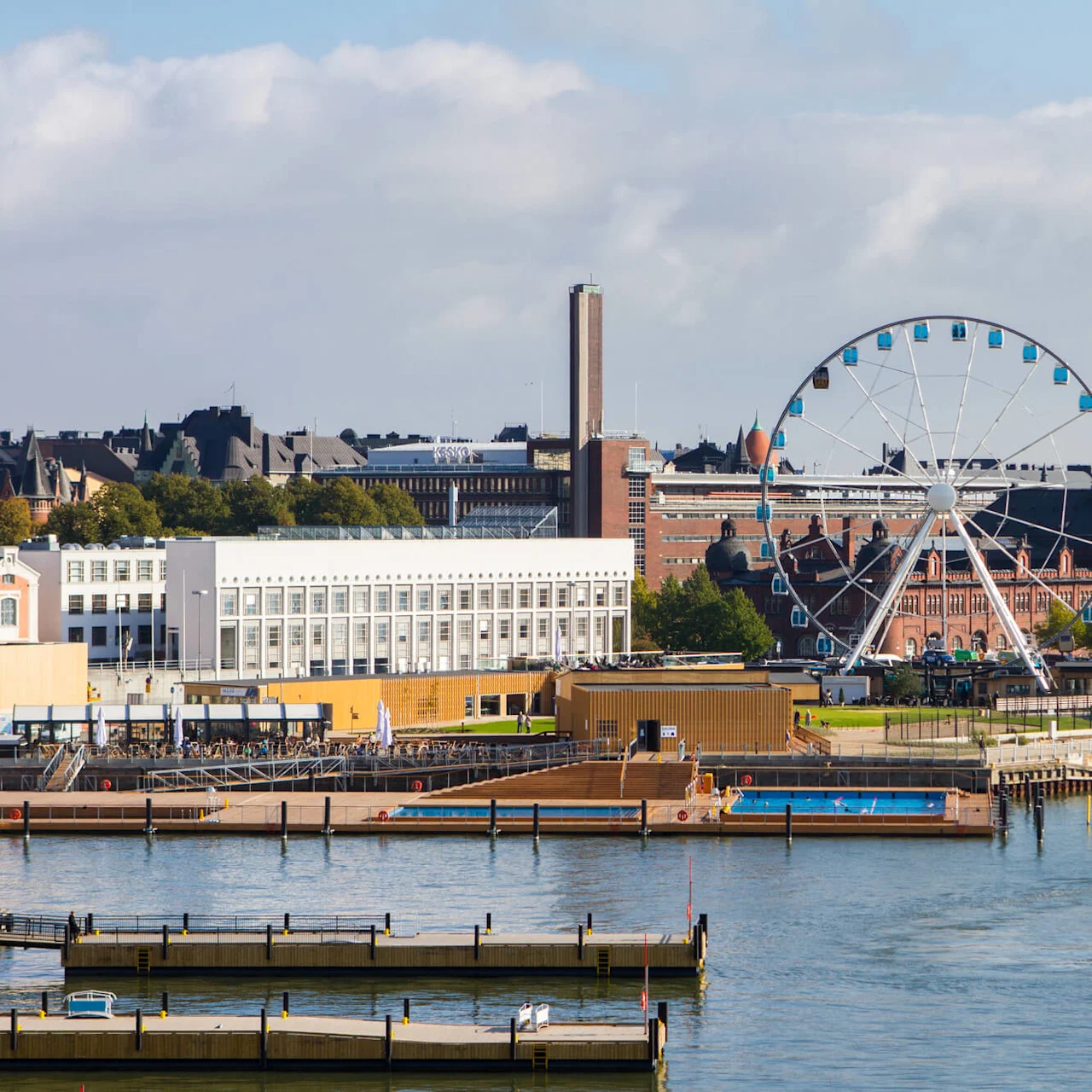 Ferris wheel stands stationary; surrounded by red-brick buildings and trees along the waterfront. Nearby, a white boat traverses calm water, with empty docks in the foreground.