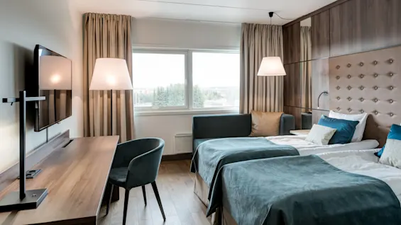 A tidy hotel room features two beds with blue covers, positioned next to a large window. A desk with a chair and wall-mounted TV complete the cozy ambiance.