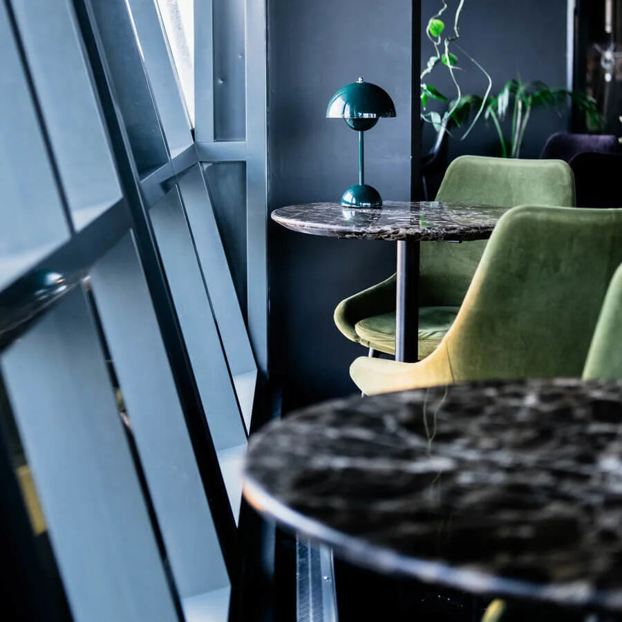 A marble-topped table supports a green lamp in a modern, dimly lit room with large, slanted windows. Green chairs and hanging plants enhance the contemporary aesthetic.