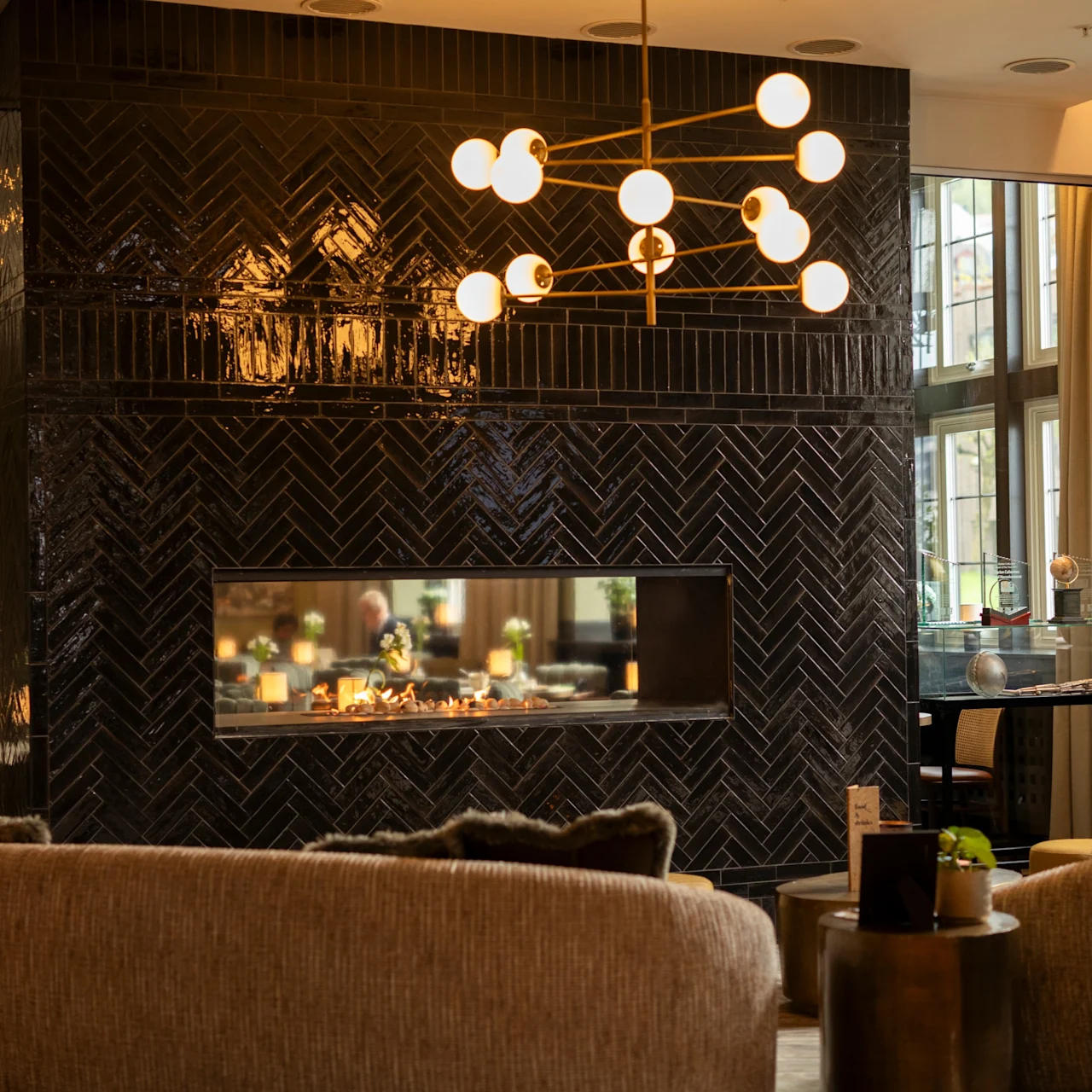 A luxurious living room features a modern fireplace set in a glossy black tile wall, illuminated by a spherical chandelier, surrounded by bookshelves and large windows.