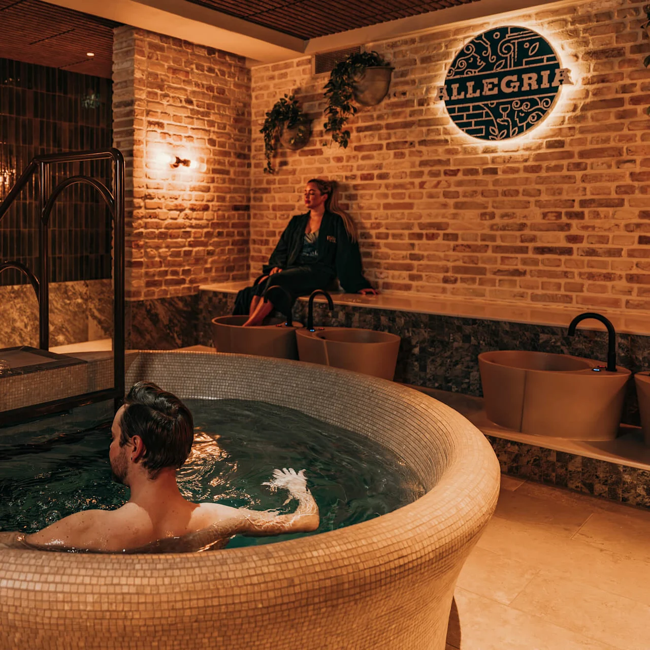 A man relaxes in a circular indoor spa pool with warm lighting, while a woman sits nearby. A sign reads "ALLEGRIA" on a brick wall.