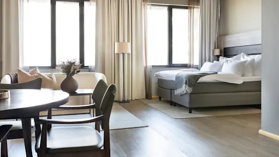 A spacious room features a neatly made bed and a cozy seating area with a vase of flowers. Sunlight streams through large windows, illuminating wooden floors and neutral-colored furniture.