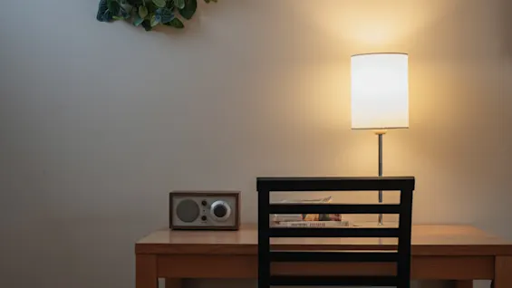 A wooden desk holds a lit lamp, a small radio, and books. A black chair is positioned in front of the desk. Greenery decorates the wall above.