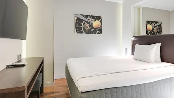 Säng med vita sängkläder i ett minimalistiskt rum med ett skrivbord i trä, väggmonterad TV och en tavla av en flygplanspropeller på väggen.