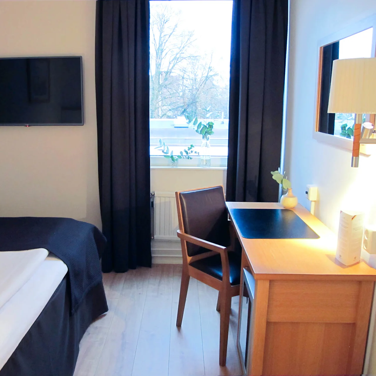 Et pent hotellrom med en seng til venstre, en veggmontert TV rett overfor og et skrivebord i tre med stol nær et vindu med mørke gardiner.