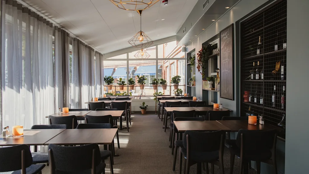 En välbelyst restaurangmatsal med flera bord redo för gäster, med stora fönster och krukväxter.