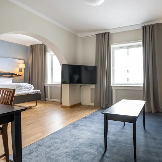 En grå sofa står i en moderne stue, ledsaget av et rektangulært kaffebord, flatskjerm-TV, lampe og stort vindu med gardiner; en seng er synlig gjennom en bue.