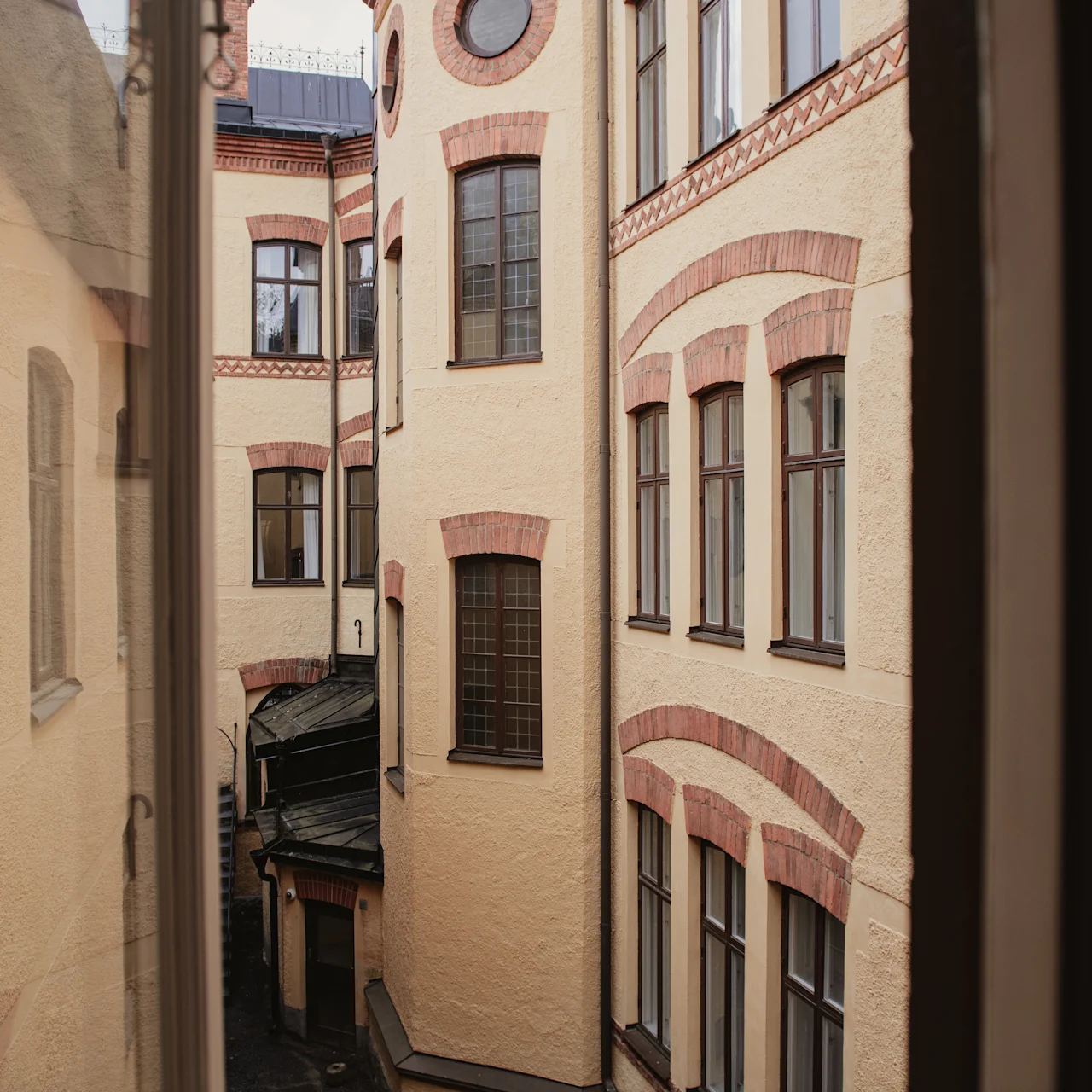 Höga, beige byggnader med röda tegelaccenter står stilla, omger en smal innergård. Flera fönster pryder väggarna, reflekterar dagsljuset. Sett från ett angränsande fönster.