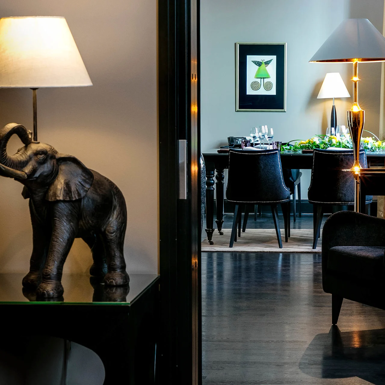 Elefant-formet lampe står på et bord, lyser opp en korridor som leder til en spisestue med et dekket bord. Et innrammet kunstverk og flere lamper pynter rommet. 