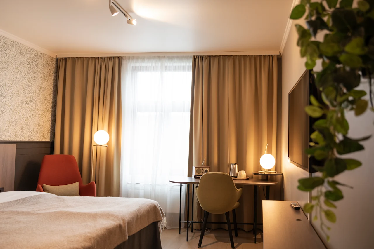Et pent organisert hotellrom har en seng og en skrivebordstol. Varm belysning lyser opp et delvis dekket vindu, med en TV og en grønn plante på veggen.