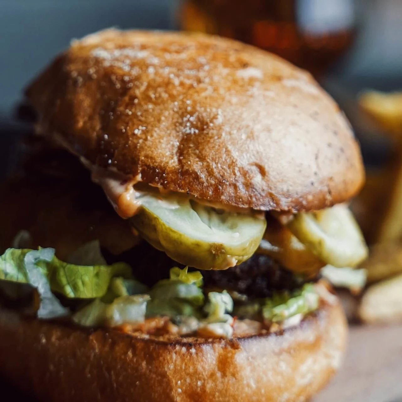 Burger med salat, pickles og saus i et ristet brød, sammen med pommes frites på en treflate i en uformell spiseomgivelse.