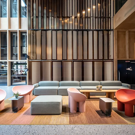 Moderne lobby med fargerike stoler og en grå sofa rundt lave bord på et fargerikt teppe, omgitt av høye trepanelskillevegger og glassvegger.