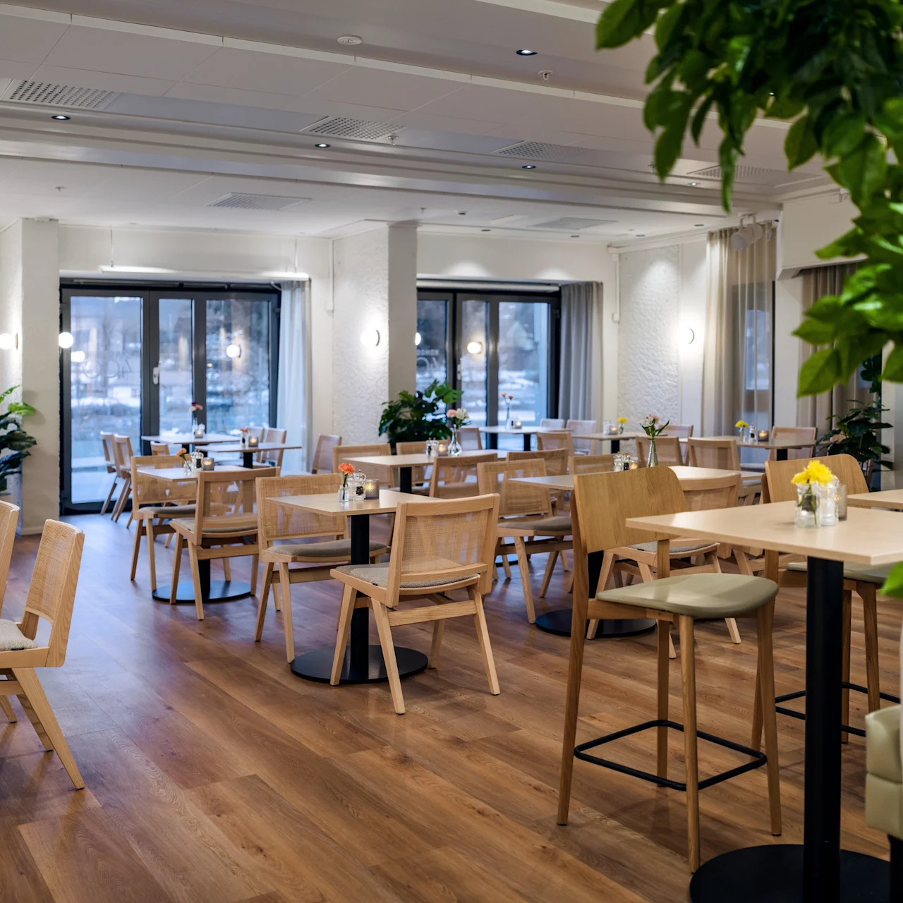 A modern dining area features wooden tables and chairs, surrounded by potted plants, under soft ceiling lights. Large windows provide natural light.