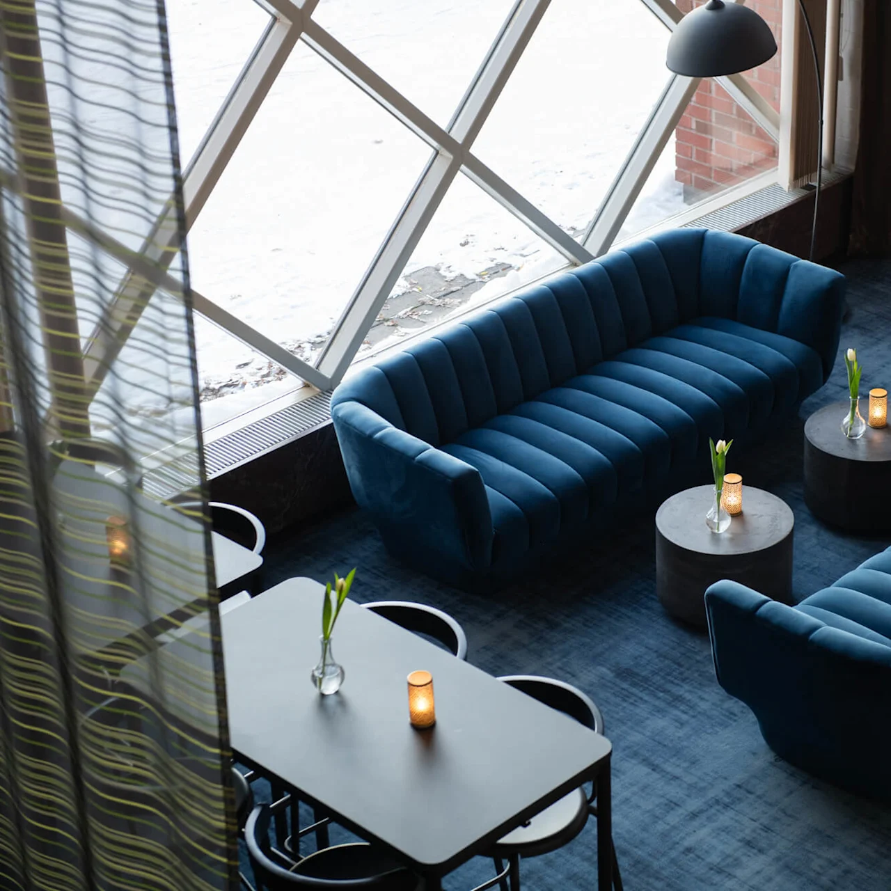 Two blue velvet sofas face each other with round tables in between, holding flowers and candles. A large window and a plant enhance the spacious, modern lounge atmosphere.