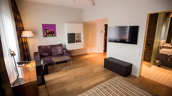 Et koselig hotellrom har en stripete seng, en sofa med salongbord, en veggmontert TV og en baderomsdør, alt på tregulv under myk belysning.