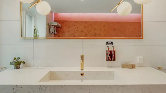 A marble bathroom counter with a sink, gold faucet, mirror, and decorative items. Ambient lighting above casts a warm glow on the scene.