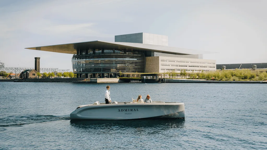 Båt märkt "Admiral Hotel" färdas med tre personer; en styr, två sitter. Omgivningen är en hamn med en stor modern byggnad med etagd design och expanderat tak.
