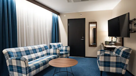 En rutig soffa och fåtölj med ett runt bord, i ett modernt, välbelyst rum med en väggmonterad tv och gardiner.