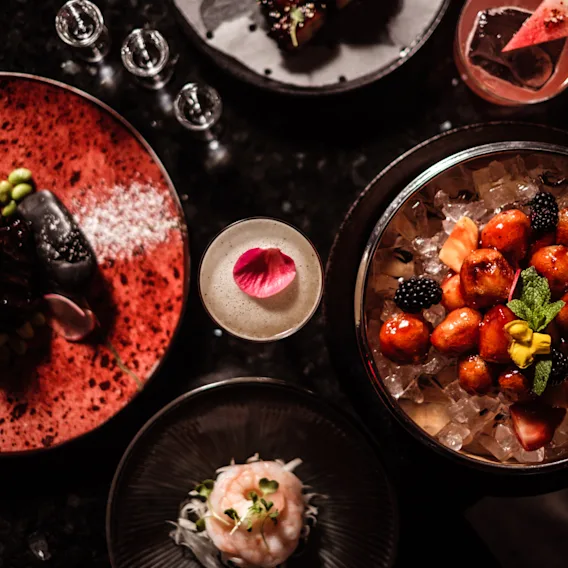 Et utvalg av gourmetretter arrangert på et mørkt bord. En cocktail med roseblad, frukt på is og ulike forretter er plassert blant elegant servise.