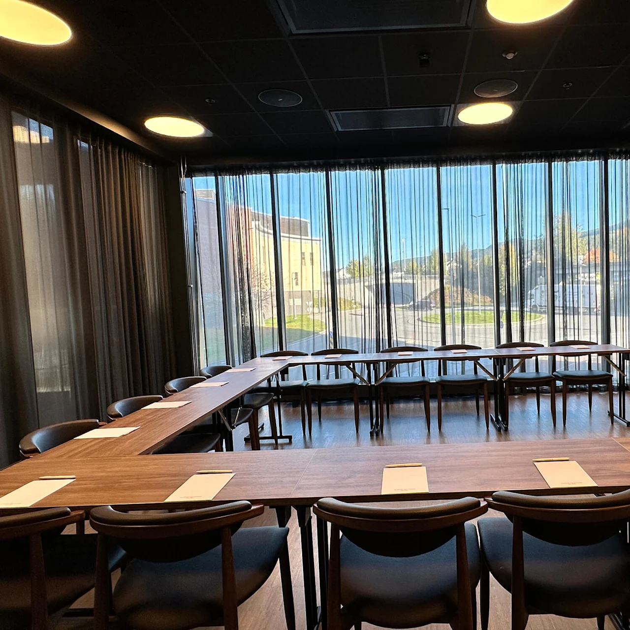 A conference room with a long table, papers and chairs, illuminated by overhead lights with large windows and curtains.