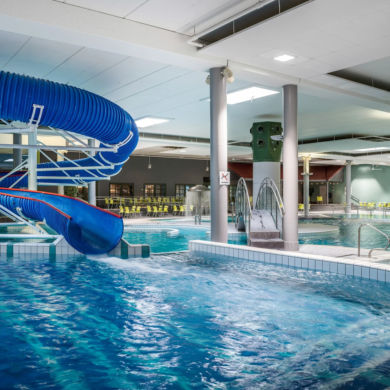 Water slide curves into an indoor swimming pool with clear blue water. Surrounding area features tiled floors, supporting columns, and lounge chairs. Bright overhead lighting illuminates the space.