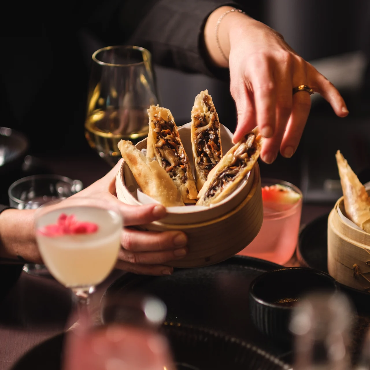 En hand hanterar en bambukorg fylld med krispiga vårrullar vid ett matbord, ackompanjerat av cocktails och olika rätter i en svagt upplyst miljö.
