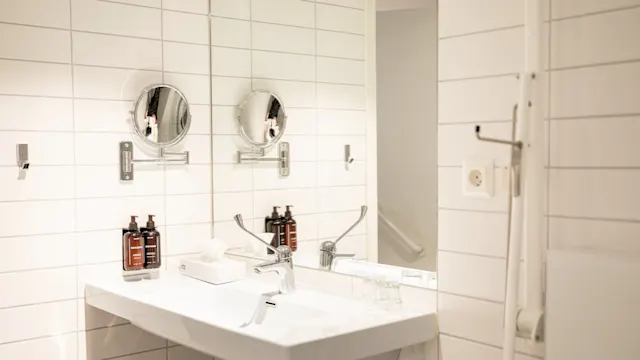 Servant flankert av speil, omgitt av hvite flisevegger; flytende såpeflasker og toiletartikler ligger ved siden av kranen på et lyst, rent bad.