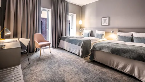 Kaksi siististi pedattua sänkyä on modernissa hotellihuoneessa, ympärillä pehmeä valaistus, pöytä, tuolit ja lattiasta kattoon ulottuvat verhot, luoden viihtyisän ja kutsuvan ilmapiirin.
