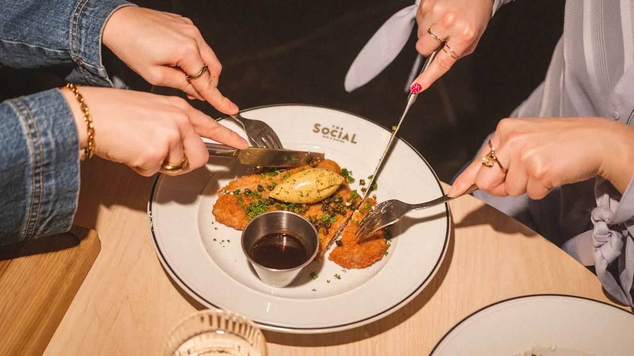 Useat kädet jakavat ja leikkaavat ateriaa lautasella, jossa näkyy sana SOCIAL, mikä viittaa yhteiseen ruokailukokemukseen.