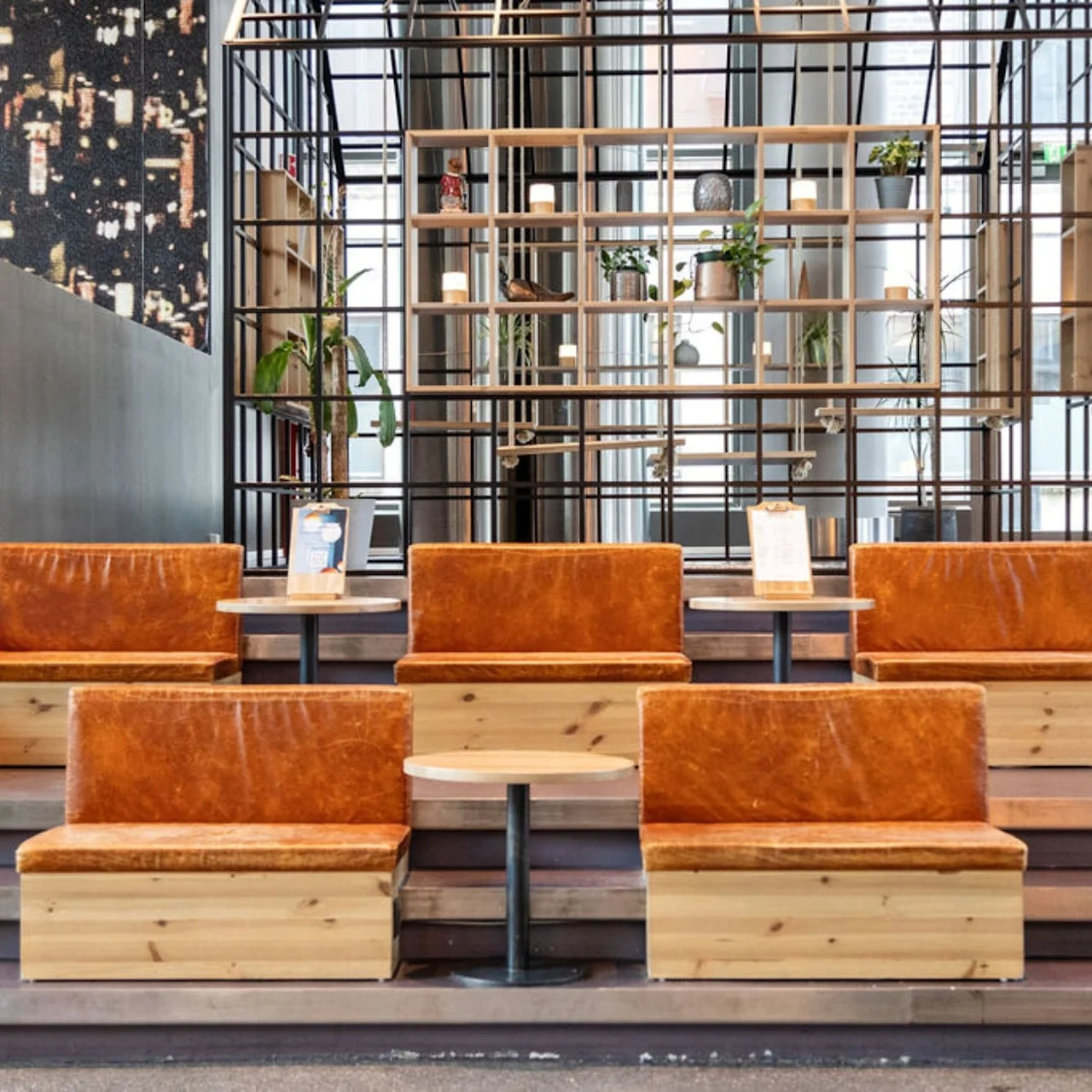 Wooden seating with leather cushions is arranged on tiered steps. Small tables accompany the seating. A decorative lattice shelf with plants stands in the background, enhancing a modern interior.