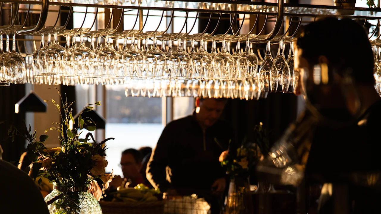 Glasses hang from a rack above a bar counter, illuminated by soft light. People gather around, with food and candles creating a warm, intimate atmosphere.