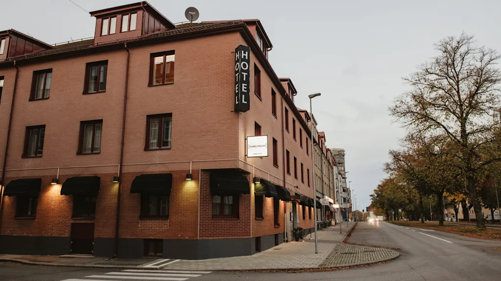 A brick hotel building on a street corner with a HOTEL sign, Home Hotel Kung Oscar. Trees line the street under an overcast sky.