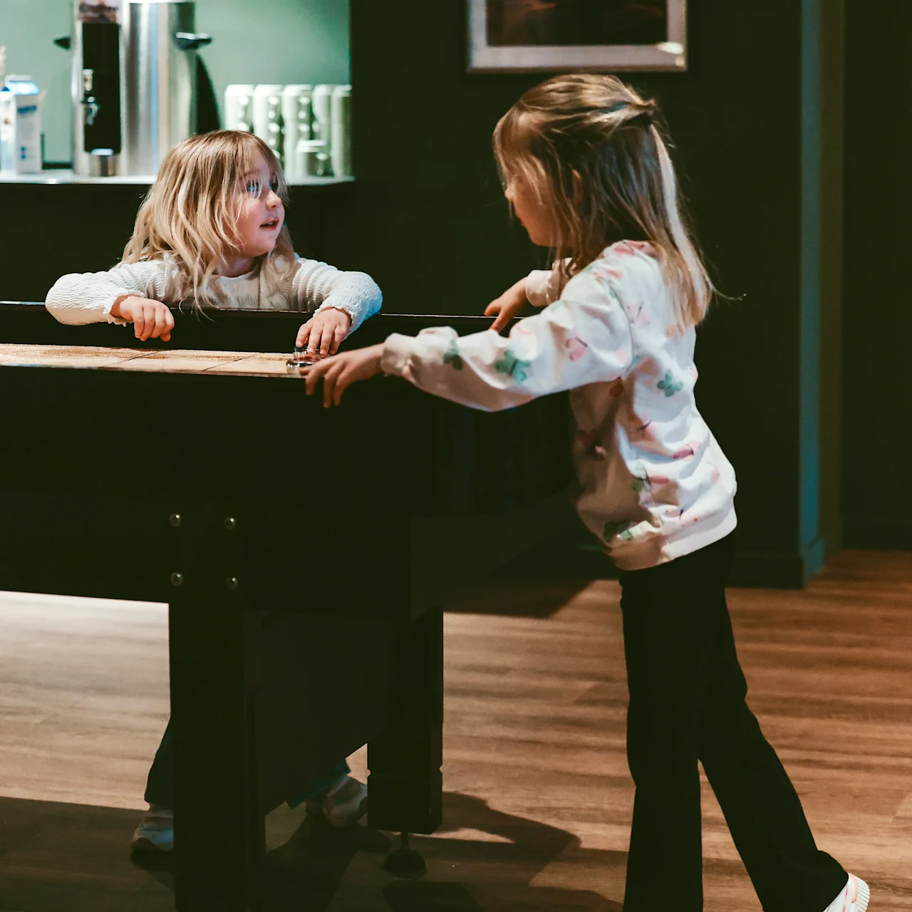 Children interact by a foosball table in a cozy room with wooden flooring. A shelf holds mugs and a pot. A framed picture hangs on the wall.