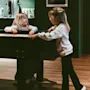 Children interact by a foosball table in a cozy room with wooden flooring. A shelf holds mugs and a pot. A framed picture hangs on the wall.