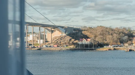 A large bridge spans a body of water, surrounded by rocky hills and trees. A building with a red roof is visible near the shoreline. The text visible is "OSMA." 