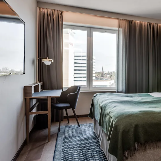 En säng med grön filt och vita kuddar är prydligt uppställd i ett hotellrum med träväggar. I närheten står ett skrivbord med en stol vid ett stort fönster.
