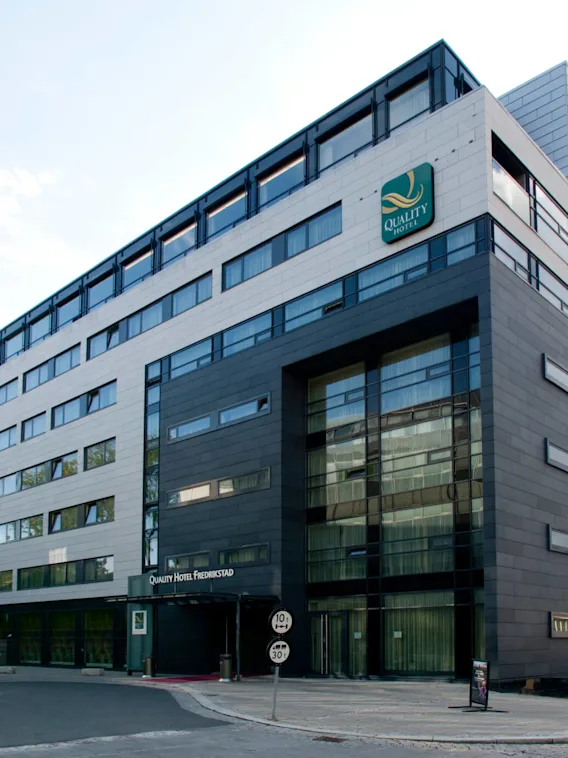 En modern hotellbyggnad med stora fönster och en fasad i grått och vitt. Text: "Quality Hotel Fredrikstad." Ligger på en lugn gata med parkerade bilar.