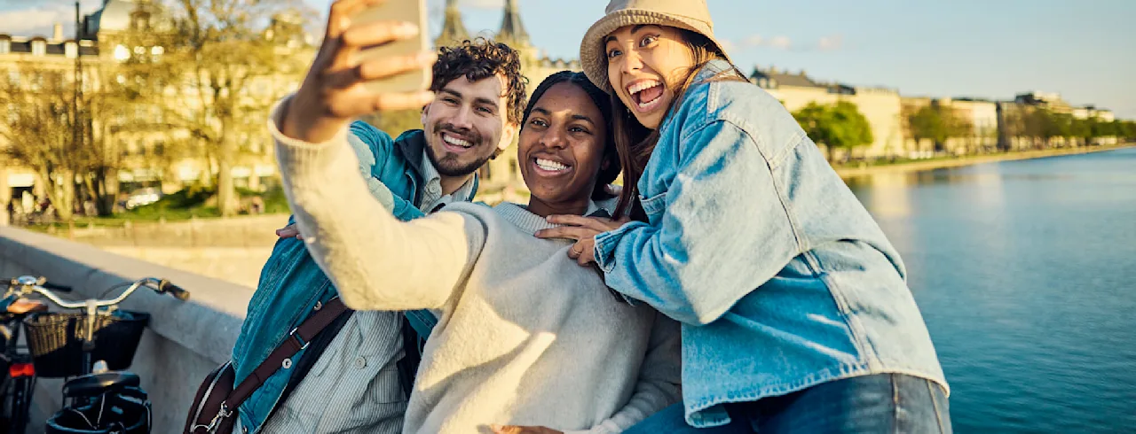 Three friends are taking a selfie by a city waterfront, smiling and embracing each other.