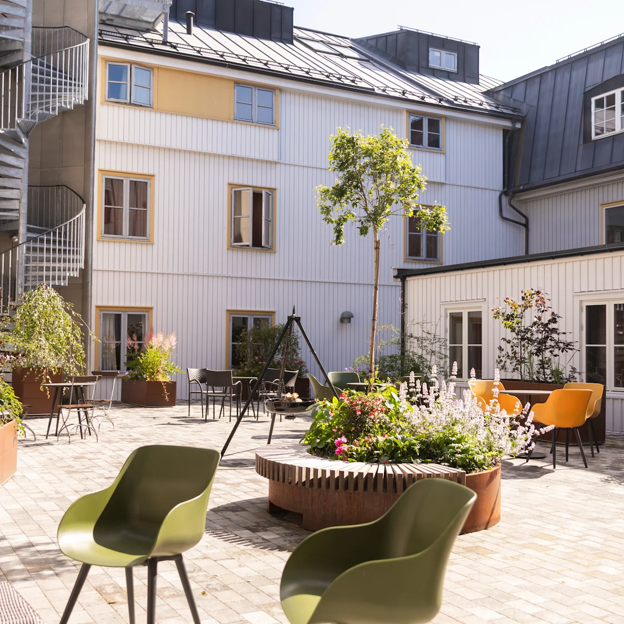 To grønne stole er placeret i en solrig gårdhave med planter, en vindeltrappe og omkringliggende hvide bygninger, hvilket skaber en fredelig udendørs atmosfære.