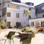 To grønne stole er placeret i en solrig gårdhave med planter, en vindeltrappe og omkringliggende hvide bygninger, hvilket skaber en fredelig udendørs atmosfære.