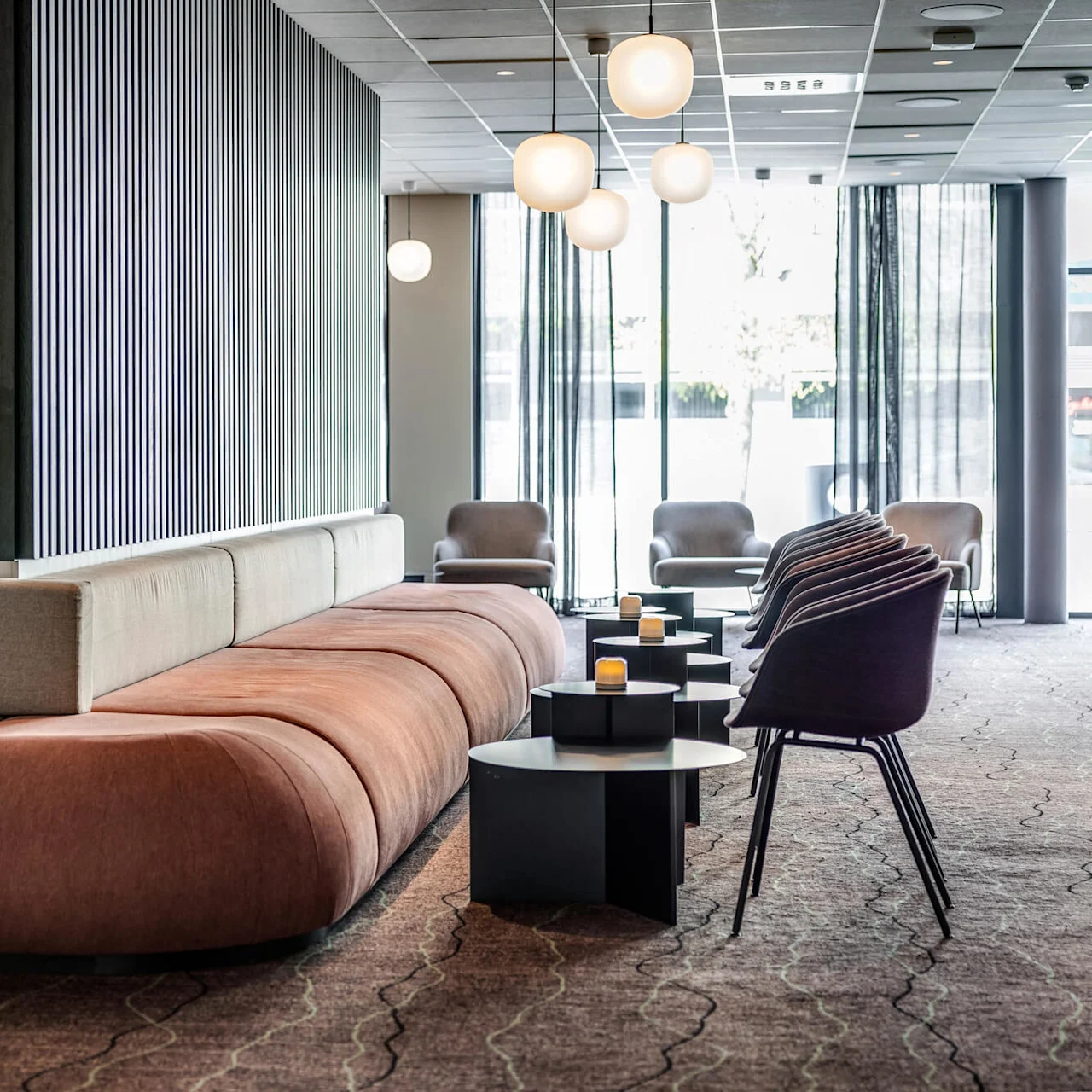 Et stort, brunt polstret siddeområde er arrangeret omkring en central skillevæg med lodrette sorte striber, omgivet af moderne stole og borde med små lys, i en lys lobby.