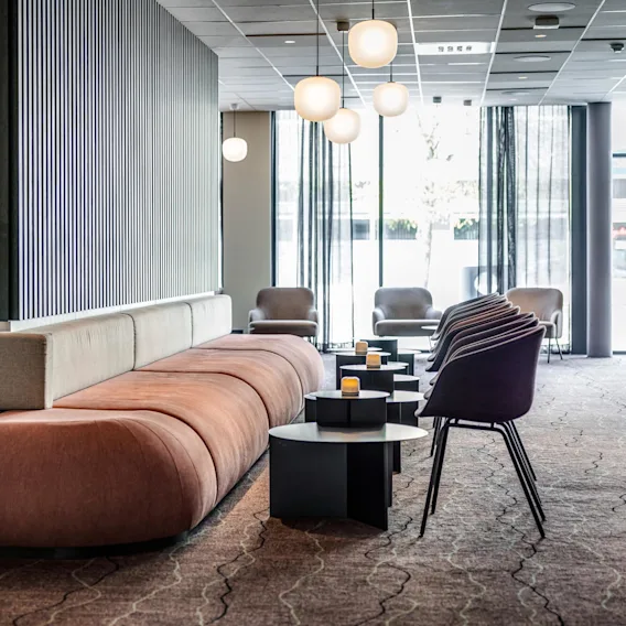 Et stort, brunt polstret sitteområde er arrangert rundt en sentral skillevegg med vertikale svarte striper, omgitt av moderne stoler og bord med små lys, i en lys lobby.