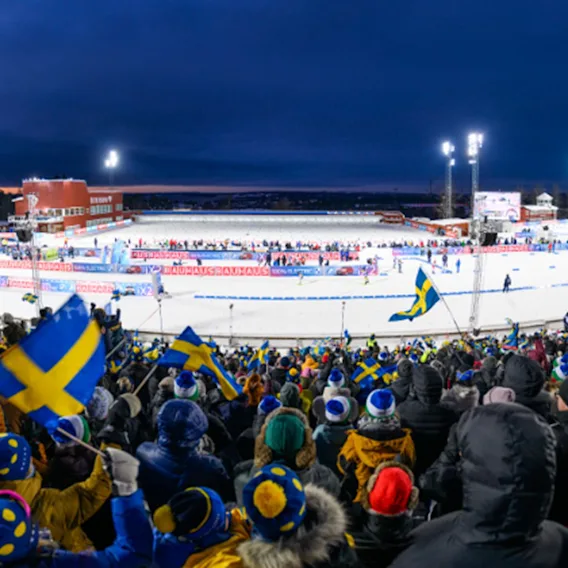 En stor folkemengde, mange viftende med flagg, ser på et vintersportsarrangement på en opplyst, snødekt bane om natten. Banner: BAUHAUS.